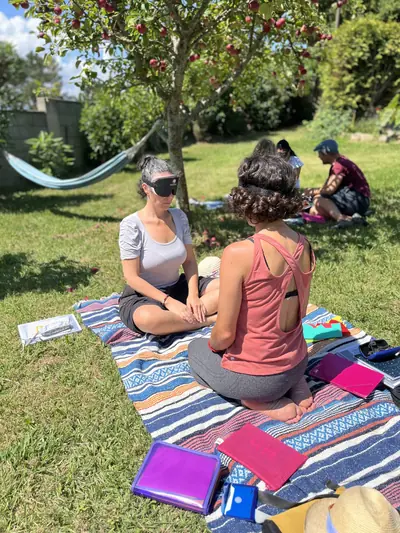 Dos participantes con antifaz practicando sentadas sobre una manta en el jardín, con más gente al fondo