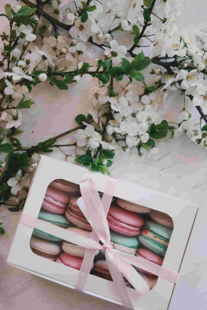 203001: La imagen muestra una composición delicada que combina flores blancas, hojas verdes y una caja blanca con dulces de colores pastel. En la parte superior e izquierda se ven varias ramas delgadas cubiertas de flores pequeñas de color blanco, con pétalos redondeados y centros ligeramente amarillentos. Entre las flores destacan hojas verdes de diferentes tonos, desde verde claro hasta verde más oscuro. En la zona inferior aparece una caja rectangular de color blanco, cerrada con una cinta rosa claro que forma un lazo suave y elegante. La tapa de la caja tiene una ventana transparente que permite ver el interior. Dentro se encuentran varios macarons de colores pastel: uno rosa claro, otro verde menta, otro beige o crema, y algunos en tonos suaves similares. El fondo general tiene colores neutros y claros, lo que hace resaltar los tonos blancos, verdes y pasteles del arreglo.
