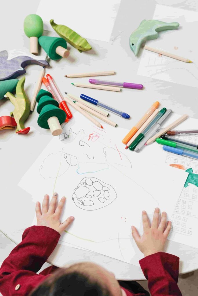 203002: 
La imagen muestra una mesa o superficie clara sobre la que hay folleos de papel con dibujos, utensilios de escritura y objetos de juguete o bloques de madera.
En la parte inferior de la imagen se ven dos manos pequeñas de un niño o niña, con la piel de tono claro y mangas de color rojo oscuro o granate. Están apoyadas sobre una hoja de papel blanca que tiene garabatos y dibujos hechos con lápices o rotuladores de diferentes colores (negro, rojo, azul y verde). Varios papeles blancos están extendidos sobre la superficie.
Alrededor de las manos hay varios lápices de madera natural (color beige claro) con puntas de colores como verde, azul y violeta. También hay rotuladores de plástico con cuerpos transparentes o gris claro y tapas de colores brillantes como naranja, verde oscuro, azul y rojo.
Hacia la parte superior izquierda de la imagen aparecen varias figuras de madera pintadas:
Una pieza de forma cónica de color verde claro y base beige.
Otra figura cónica en verde más intenso con base beige.
Una pieza que parece una vaina o forma orgánica en verde claro con detalles más oscuros.
Otras piezas que parecen siluetas de animales o formas con tonos verde pastel y verde agua.
Los colores dominantes en la imagen son: Blanco del papel y fondo. Verde en diferentes tonos (desde verde menta hasta verde oscuro) en las figuras de madera. Beige claro o madera natural en los lápices. Rojo/granate en las mangas del suéter del niño/a. Colores vivos en los rotuladores y puntas de lápices (naranja, azul, violeta, verde).
La composición sugiere un momento de dibujo o juego creativo, con objetos de arte y juguetes dispersos sobre una superficie iluminada y en tonos mayormente claros y pasteles.
