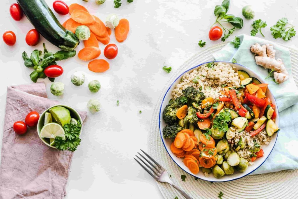 203005:
La imagen muestra una escena culinaria con vegetales frescos y un plato preparado colocado sobre una superficie clara, probablemente una mesa de color blanco o muy claro.A la derecha hay un plato redondo con borde azul oscuro que contiene una mezcla de alimentos de colores vibrantes: Brócoli de color verde intenso. Zanahorias en rodajas de color naranja brillante. Pimientos rojos en tiras o trozos de rojo vivo. Trozos de calabacín verde claro y oscuro. Granos cocidos de color beige claro, que parecen ser quinoa o algún cereal similar. Hojas pequeñas de perejil verde, esparcidas encima de los vegetales.
A la izquierda del plato, dispersos sobre la superficie, hay varias verduras y condimentos: Tomates cherry de color rojo brillante. Coles de Bruselas de color verde pálido con toques más claros. Rodajas de zanahoria extra de color naranja. Un calabacín grande de color verde oscuro apoyado en ángulo. Un pequeño cuenco verde claro que contiene gajos de lima (verde y amarillo pálido) y hojas de perejil de color verde vivo.
Debajo del cuenco hay una servilleta o paño de tela de color rosa pálido o malva claro. En la parte inferior derecha, hay un tenedor metálico plateado colocado sobre la mesa. Cerca del plato se ve otra servilleta de color verde agua o verde muy claro, que añade un toque de color suave al conjunto.