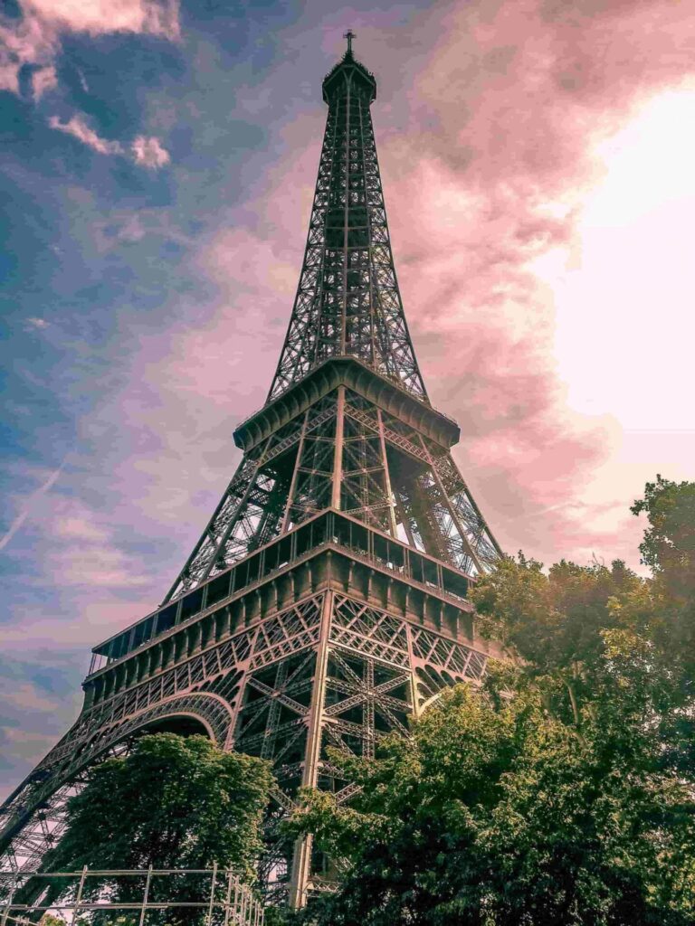 203015:
Esta es una toma de ángulo contrapicado extremo que presenta la Torre Eiffel, una estructura de hierro marrón oscuro o gris oscuro de forma cónica y compleja red de celosías, que domina la composición al extenderse verticalmente desde la base hasta la parte superior del cuadro. El ángulo acentúa sus líneas diagonales y su monumentalidad. En el primer plano y a los lados, las copas de árboles frondosos de color verde oscuro y profundo tienen formas orgánicas y redondeadas que enmarcan la base de la estructura metálica. El cielo de fondo es vibrante, iluminado por un atardecer que lo tiñe de tonos intensos de rosa, malva y azul claro, con grandes nubes que transicionan de colores más claros y brillantes a tonos más oscuros.