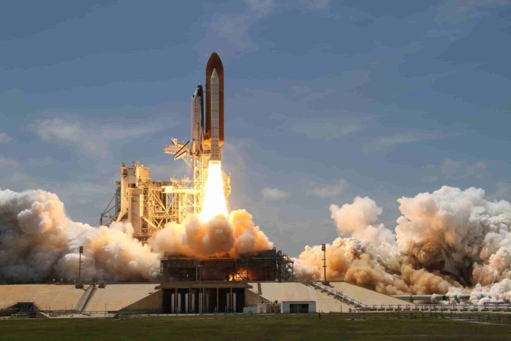 203016:
La imagen es un plano abierto que inmortaliza el dramático ascenso vertical de un transbordador espacial, envuelto en el humo y el fuego de sus propulsores. La nave espacial se ubica en el centro superior, mostrando su forma alargada y cilíndrica: el gran tanque externo es de un tono naranja rojizo y está flanqueado por los propulsores laterales de color blanco puro. El elemento más dinámico es la columna de propulsión que nace en la base: es una masa de fuego incandescente de color blanco y amarillo brillante que se expande hacia abajo en una forma cónica invertida. Este fuego está rodeado por grandes nubes de humo y vapor turbulento de tonalidades que van del marrón anaranjado denso al blanco puro, extendiéndose lateralmente sobre la plataforma. La estructura de lanzamiento se aprecia como un conjunto de líneas verticales y horizontales de color gris claro y beige en la base del cohete. Todo esto ocurre bajo un cielo de azul claro parcialmente cubierto por nubes blancas dispersas.